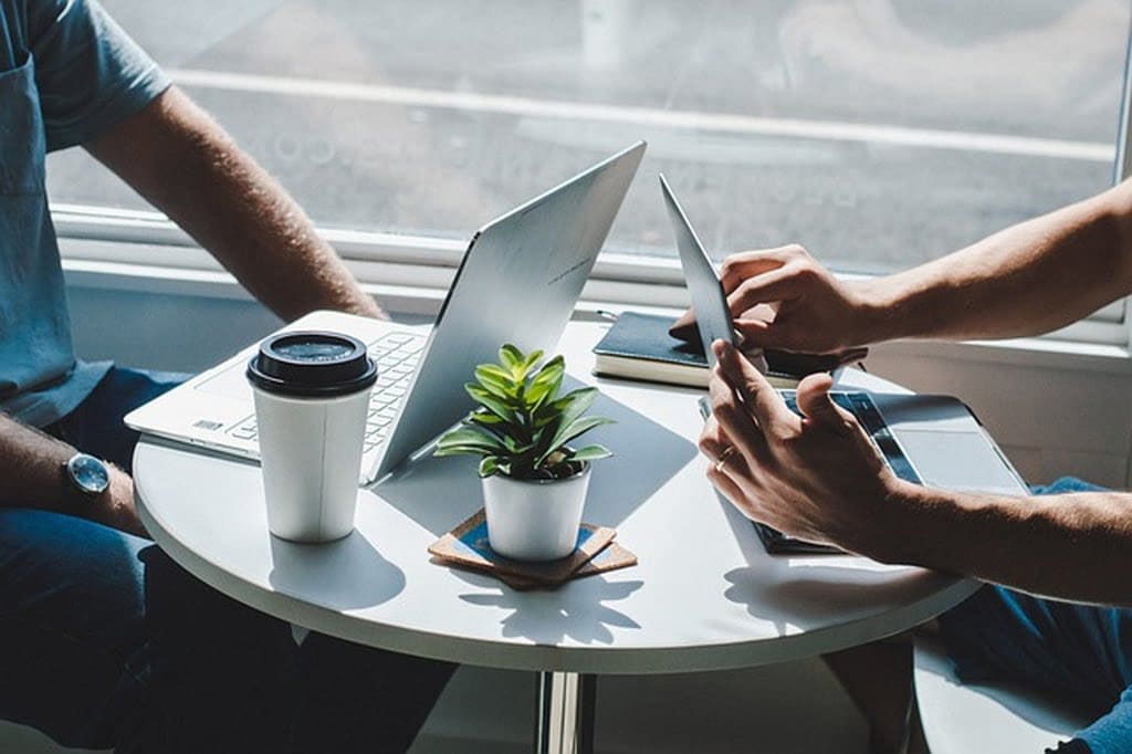 Duas pessoas à mesa num ambiente comercial. Sobre a mesa está um pequeno vaso de plantas, um copo de café com tampa, um caderno de anotações, e dispositivos como notebook e tablet. As pessoas simbolizam estar fazendo controle de aluguel via software de controle